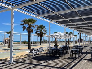 An outdoor seating area with tables and chairs, a covered walkway, and palm trees. - Apartment with AC, port, pets welcome - 4 guests (Port Leucate)
