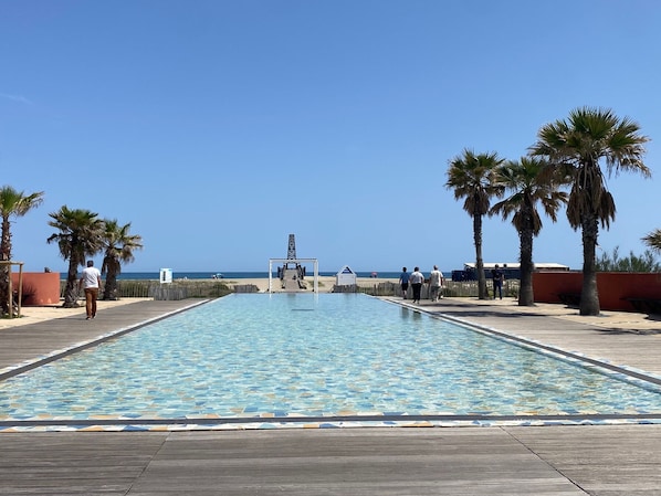 A swimming pool with a tiled pattern, surrounded by a wooden deck, palm trees, and a clear blue sky. - Apartment with AC, port, pets welcome - 4 guests (Port Leucate)