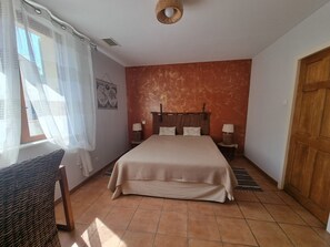 Ein Schlafzimmer mit einem Bett, zwei Nachttischen, einem Sessel, einer Holztür und einem Fenster mit Vorhängen.