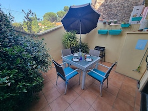 Eine Terrasse mit Tisch für zwei Personen, Stühlen, einem blauen Sonnenschirm und Blumentöpfen.