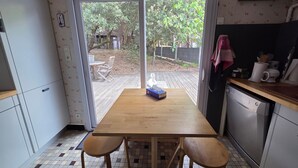 Een keuken met een houten tafel en twee stoelen, een vaatwasser, en uitzicht op een achtertuin met bomen en een houten terras.