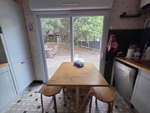 Eine Küche mit Holztisch und zwei Stühlen, einem Geschirrspüler sowie Blick auf einen Garten mit Bäumen und einer Holzterrasse.