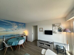 A living room with a painting of a seascape, a television, and a dining area with a table and chairs. - Apartment with parking - 4 guests. (Cabourg)