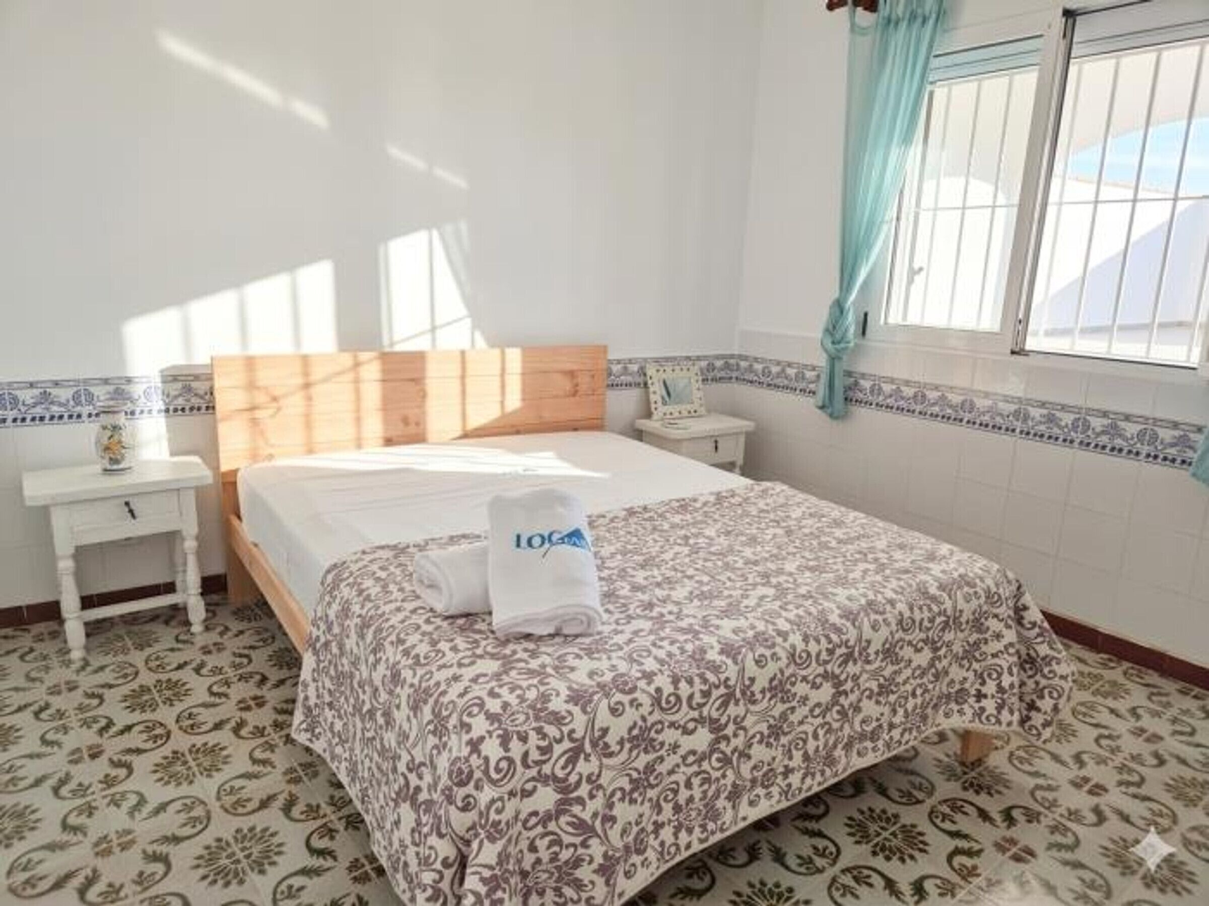 A bedroom with a bed, a nightstand, a window with curtains, and patterned floor tiles.