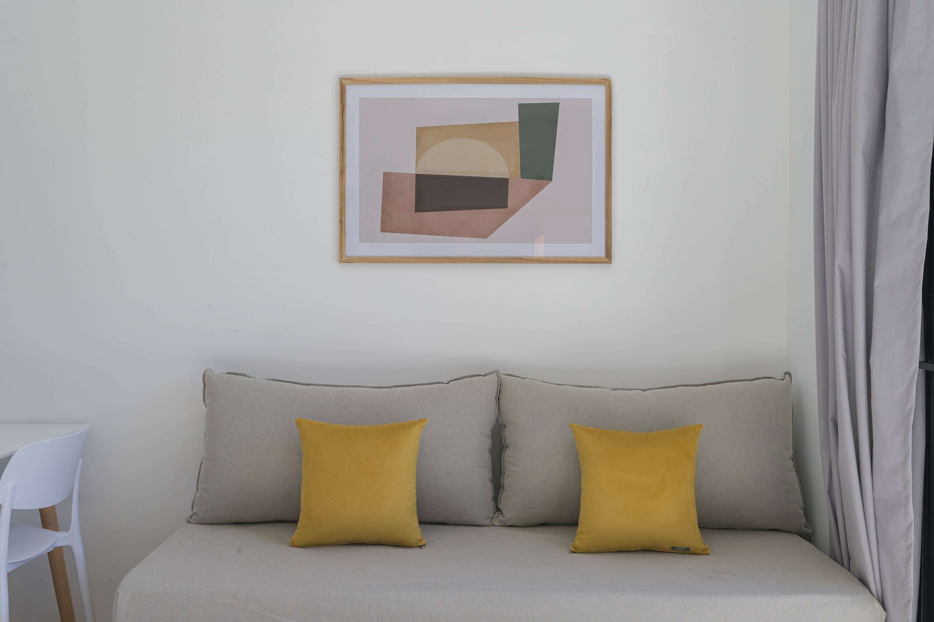 Un canapé aux couleurs neutres agrémenté de deux coussins, une œuvre d’art abstraite encadrée accrochée au mur, et une chaise blanche placée à gauche.