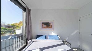 Et soveværelse med et stort vindue, en seng med hvide sengetøj, et natbord med en lampe, og et abstrakt kunstværk i ramme på væggen.