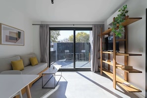 A modern living room with a sofa, a table, a bookshelf, and a sliding glass door leading to an outdoor area. - MOT Rentals + Black Ravignani 2C - 2 Bd Palermo Hollywood (Buenos Aires)