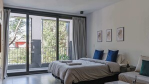 Ein Schlafzimmer mit einer großen Glaswand, die auf einen Balkon führt, einem Bett mit grauer Bettdecke, einem Nachttisch mit Lampe und drei gerahmten Bildern an der Wand.