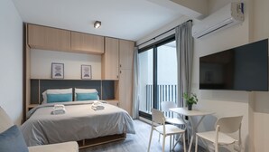 Una habitación de hotel moderna con una cama, mesitas de noche, un televisor, un balcón con cortinas y una pequeña mesa con sillas.