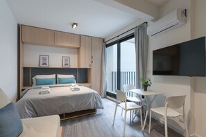 Ein modernes Hotelzimmer mit Bett, Nachttischen, einem Fernseher, einem Balkon mit Vorhängen und einem kleinen Tisch mit Stühlen.