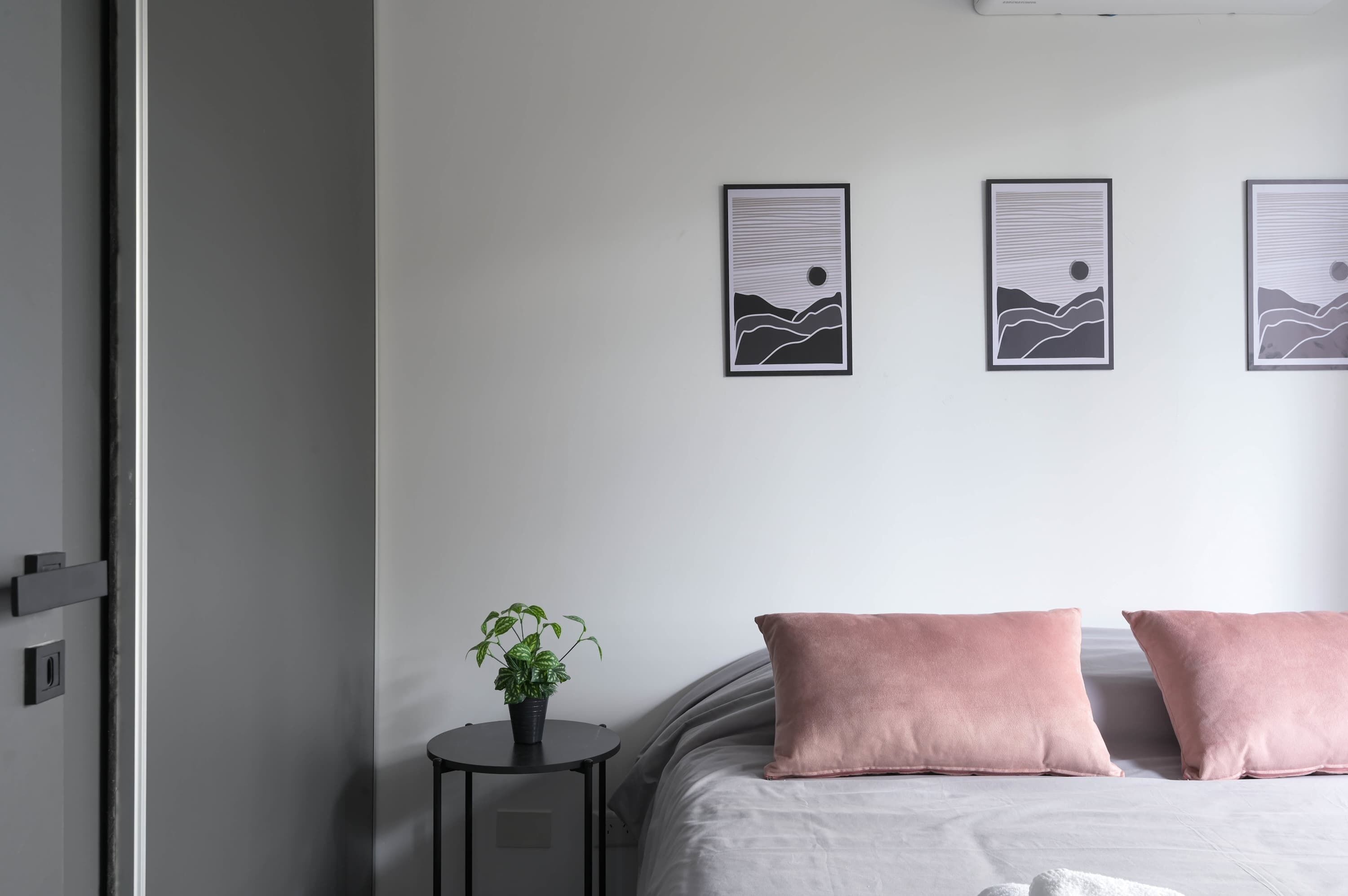 Une chambre à coucher avec un lit, deux oreillers, une table de chevet ornée d’une plante et trois tableaux encadrés accrochés au mur.