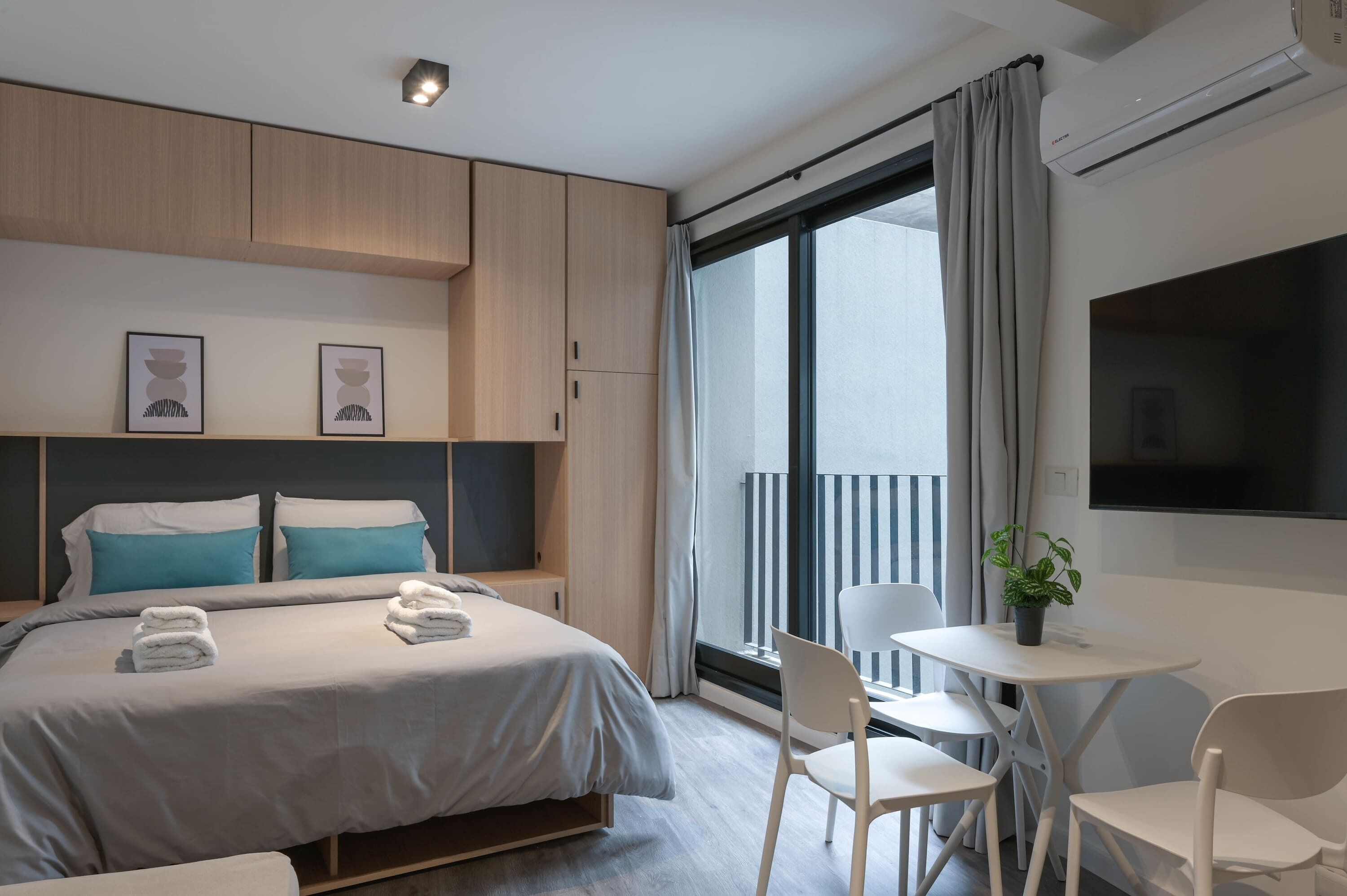 Una habitación de hotel moderna con una cama grande, una pequeña mesa con sillas, un televisor y un balcón con cortinas.