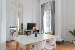 Ein Essbereich mit einem weißen Tisch, Stühlen, einem Fernseher und einer Vase mit Blumen.