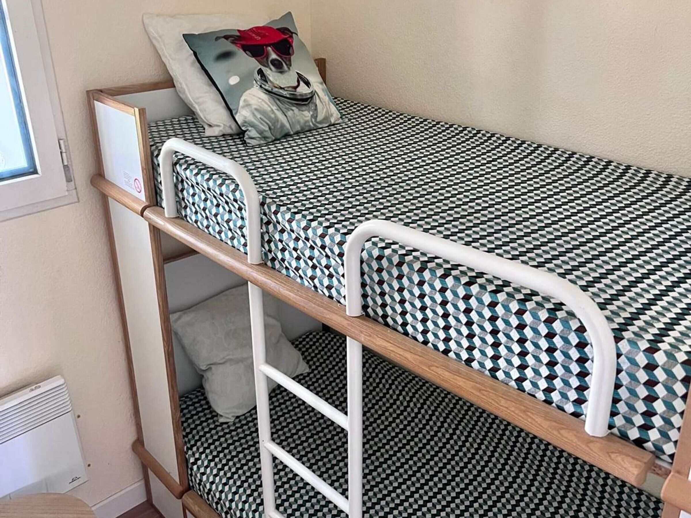 A bunk bed with a checkered mattress, a pillow with a dog illustration, and a window with blinds.