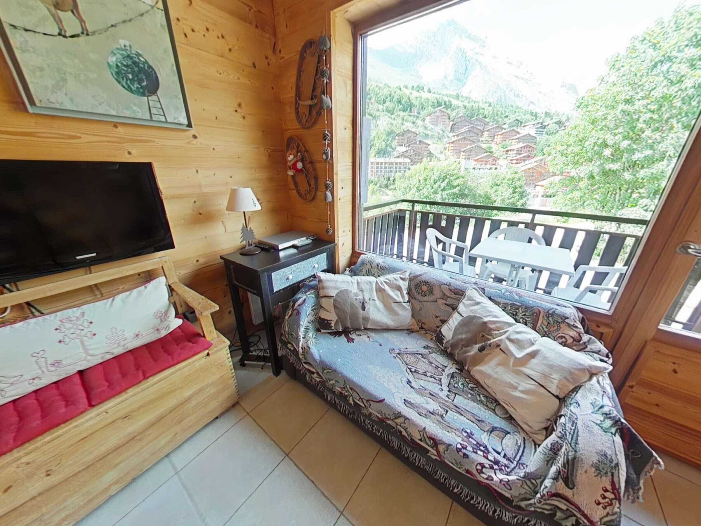 Ein gemütliches Zimmer mit Bett, Fernseher, kleinem Tisch mit Lampe und Balkon mit Bergblick.