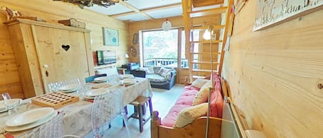 A cozy cabin interior with a dining area, a sofa, a television, and a wooden ceiling.