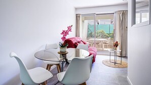 A modern living room with a glass table, white chairs, a red sofa, and a view of a garden through a sliding door. - Modern apartment, AC and parking - 3 guests (Canet-en-Roussillon)