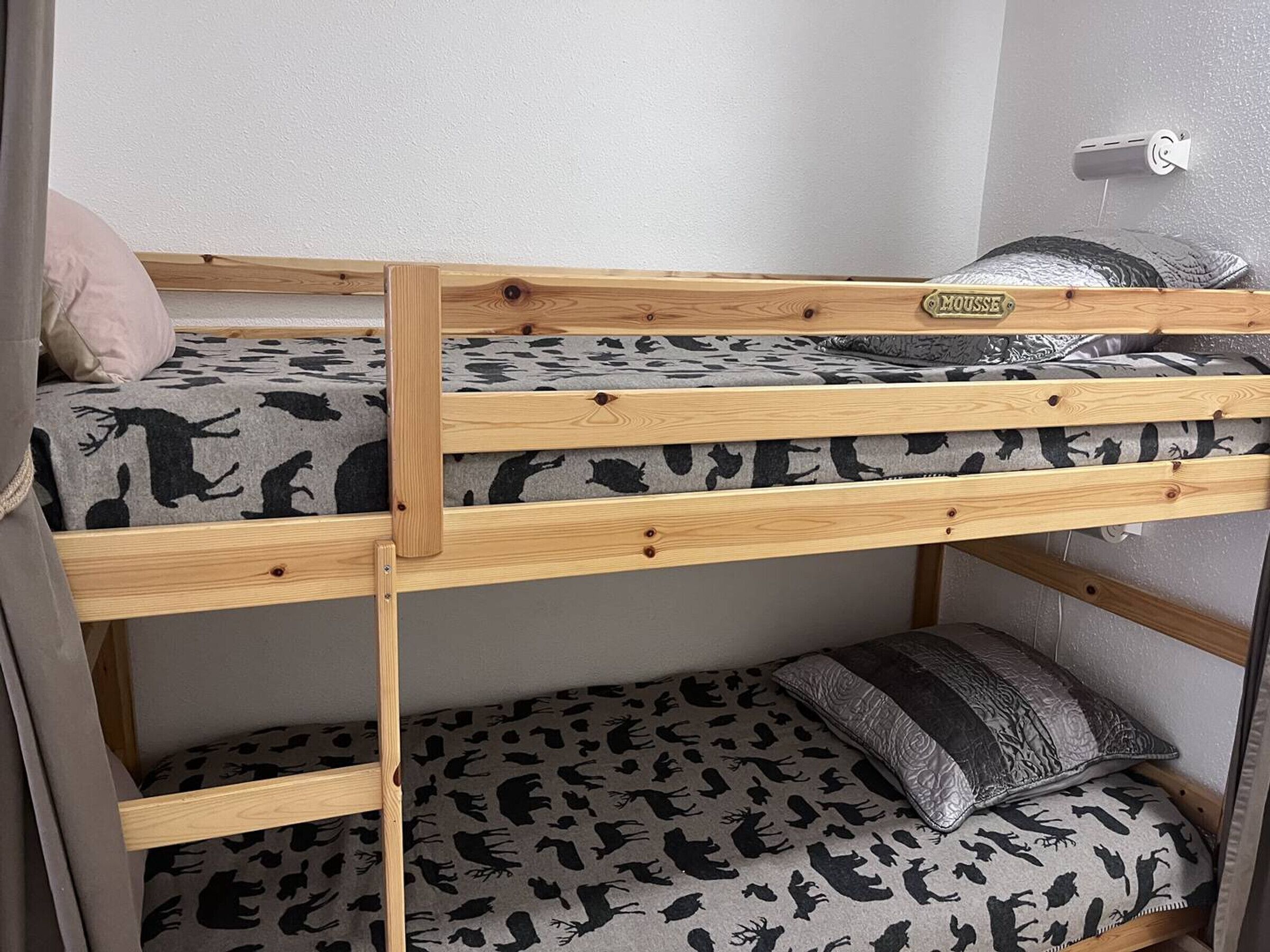 Un lit superposé avec une étiquette indiquant « MOUSSE », un oreiller et un luminaire fixé au mur.