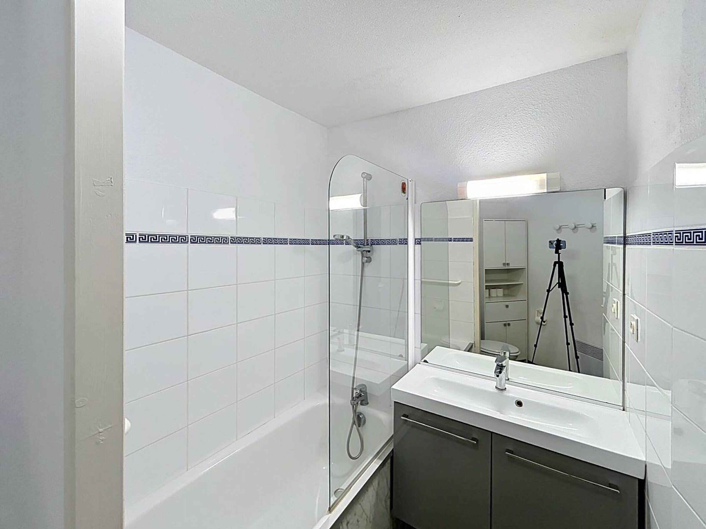 Uma casa de banho moderna com azulejos brancos, cabine de duche em vidro, toucador com espelho e uma câmara montada em tripé.