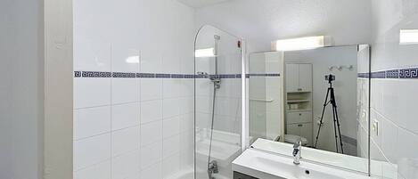 Uma casa de banho moderna com azulejos brancos, cabine de duche em vidro, toucador com espelho e uma câmara montada em tripé.