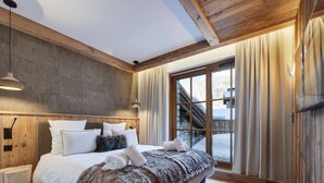 Ett sovrum med en stor säng, ett träbeklätt sänggavel, sänglampor, utsikt över ett snötäckt landskap genom ett fönster och ett träpanelat tak.