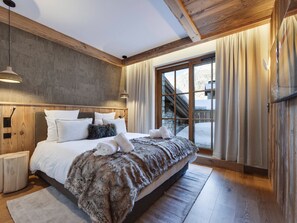 A bedroom with a large bed, wooden headboard, bedside lamps, a view of a snowy landscape through a window, and a wooden ceiling. - Apartment mountain view - 8 guests (Val-d'Isère)