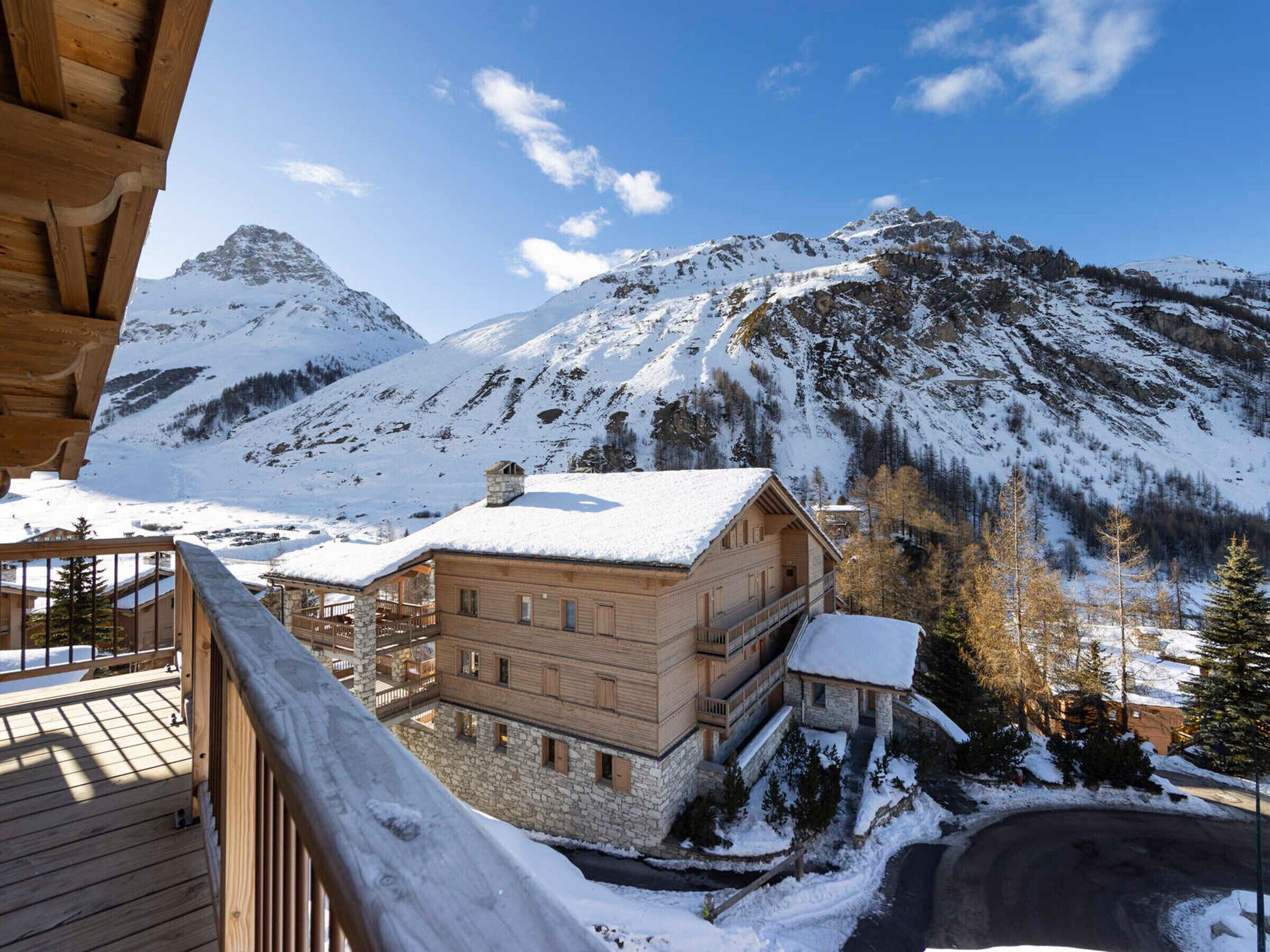 A wooden chalet with a stone foundation, a balcony, and a view of snow-covered mountains.