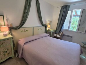 Ein Schlafzimmer mit einem Bett, einem Nachttisch mit Lampe, einem Sessel, einem Schreibtisch mit Lampe und einem Fenster mit Vorhängen.