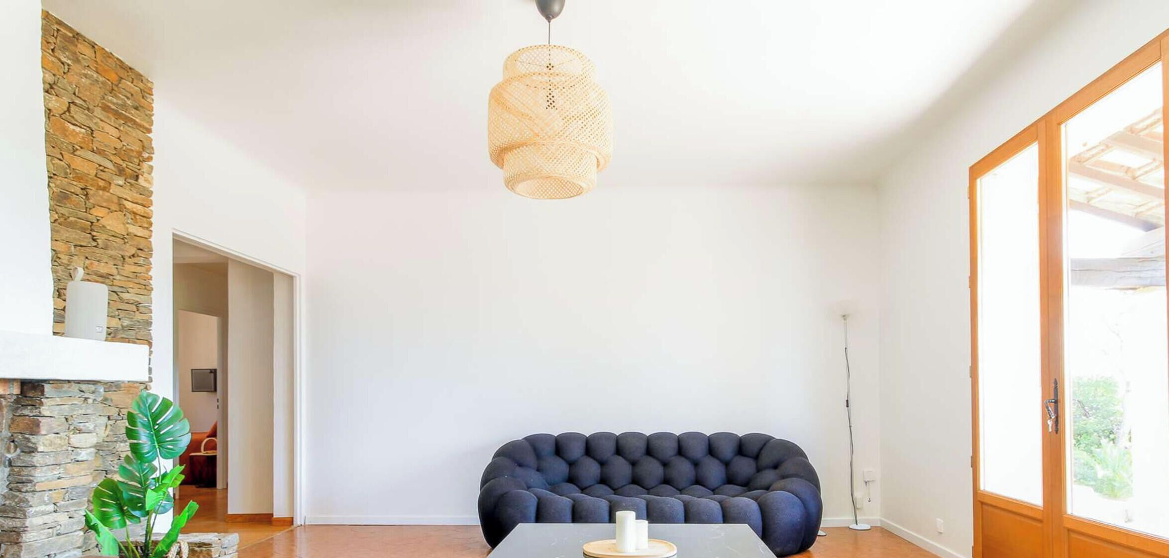 Una sala de estar moderna con pared de piedra, un sofá tapizado oscuro, una lámpara colgante y vistas a través de una puerta de cristal.