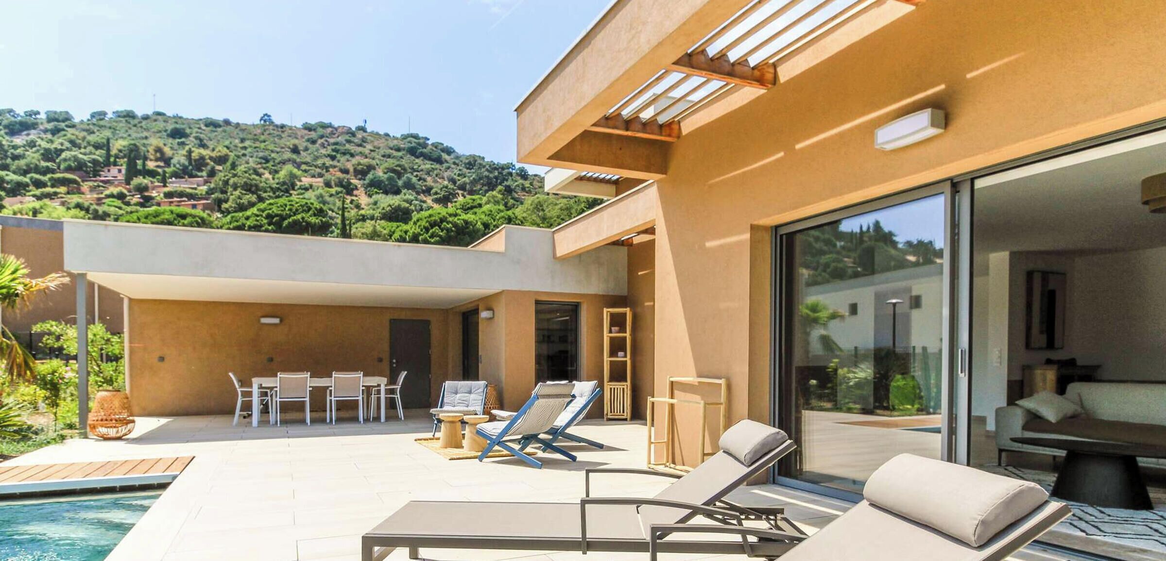 A modern outdoor pool area with lounge chairs, a dining set, and a view of a hillside.