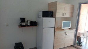 A compact kitchen with a microwave, coffee maker, and a small refrigerator. There is a wooden cabinet with a TV on top, and a trash bin is visible. - Studio cabin sea view - 3 guests (Cavalaire-sur-Mer)