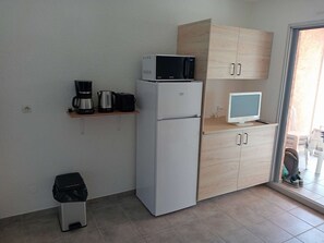 Eine kleine Küche mit Mikrowelle, Kaffeemaschine und einem kleinen Kühlschrank. Ein Holzschrank mit Fernseher darauf ist zu sehen, und ein Mülleimer steht dort.