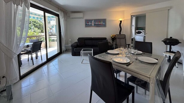 A dining area with a table set for four, a sofa, a painting, and a kitchenette. - Apartment with AC, terrace - 4 guests (Cavalaire-sur-Mer)