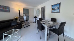A modern living room with a dining table set for four, a sofa, a coffee table, and a kitchen in the background. - Apartment with AC, terrace - 4 guests (Cavalaire-sur-Mer)