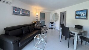A modern living room with a black sofa, a dining table with chairs, a small white stool, a floor lamp, and a painting on the wall. - Apartment with AC, terrace - 4 guests (Cavalaire-sur-Mer)