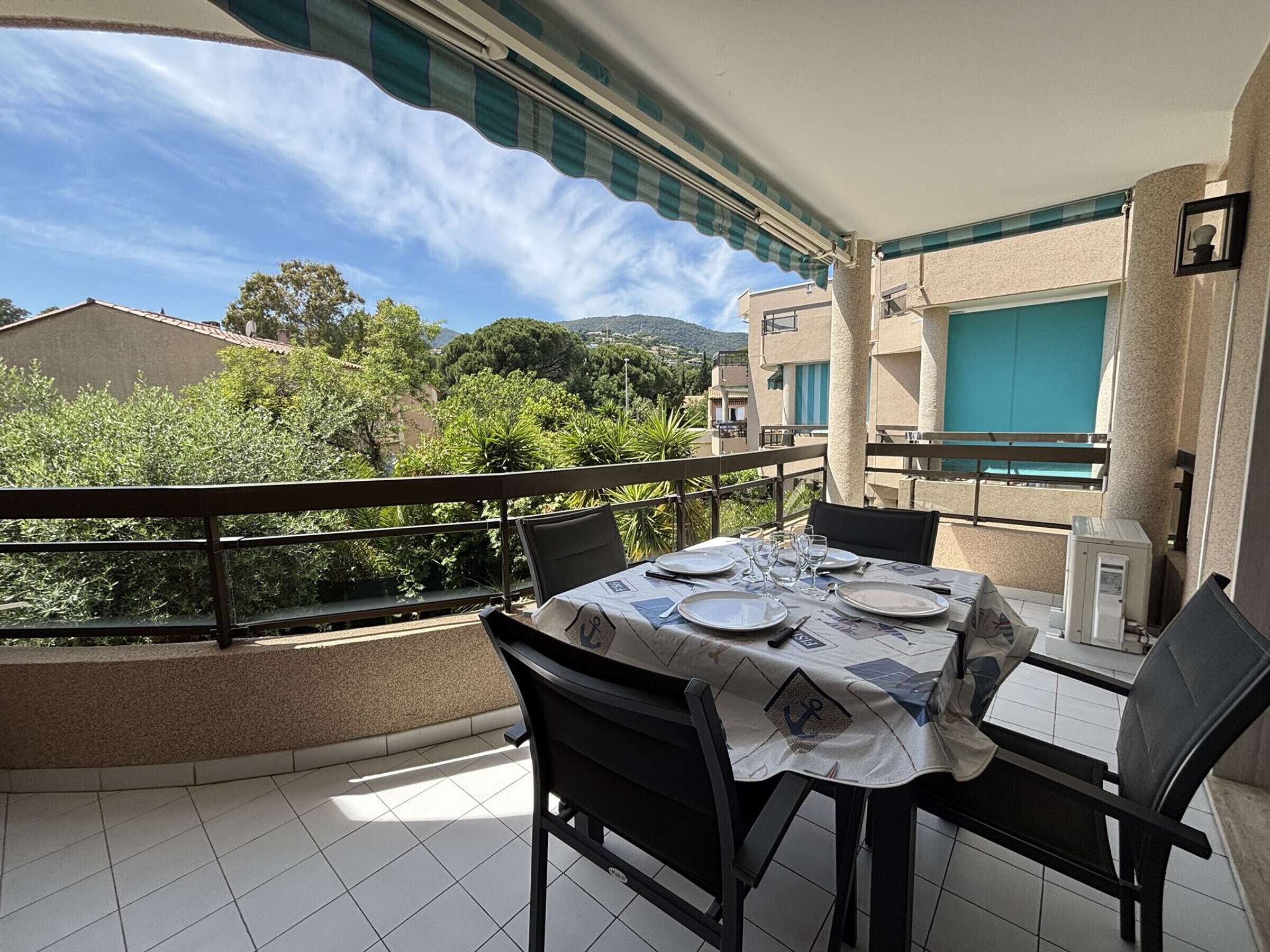 A balcony with a table set for dining, chairs, a small table lamp, and a view of greenery and buildings.