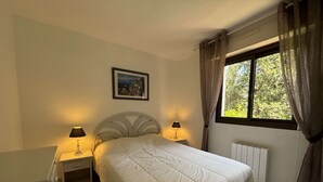 A bedroom with a bed, two bedside lamps, a radiator, a window with curtains, and a framed picture on the wall. - Apartment with AC, terrace - 4 guests (Cavalaire-sur-Mer)