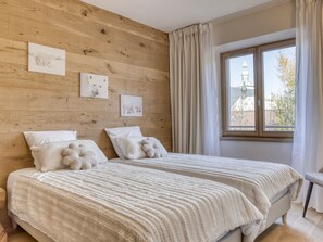 Ein Schlafzimmer mit hölzernem Kopfteil, zwei Betten mit weißer Bettwäsche, einem Fenster mit Vorhängen und Wandbildern.
