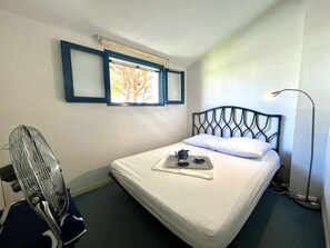Un lit simple avec une tête de lit en métal, une petite fenêtre avec des stores, une lampe sur pied et un ventilateur.