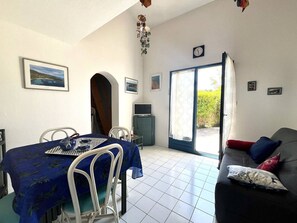 Un coin repas avec une table, des chaises et une nappe bleue. On aperçoit un canapé avec des coussins et un téléviseur au fond.
