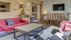 Een gezellige woonkamer met een houten wand, een bank, een salontafel, een televisie en een eethoek met stoelen.