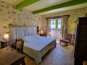 Ein Schlafzimmer mit Bett, Sessel, Tisch, Lampe, einem Fenster mit Vorhängen und einer Tür.