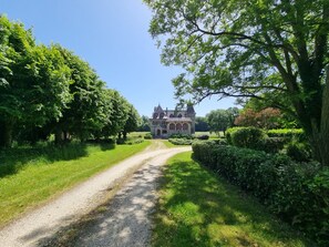 Ein Kiesweg führt zu einem großen, historischen Gebäude mit einem markanten Turm, umgeben von üppigem Grün und Bäumen.