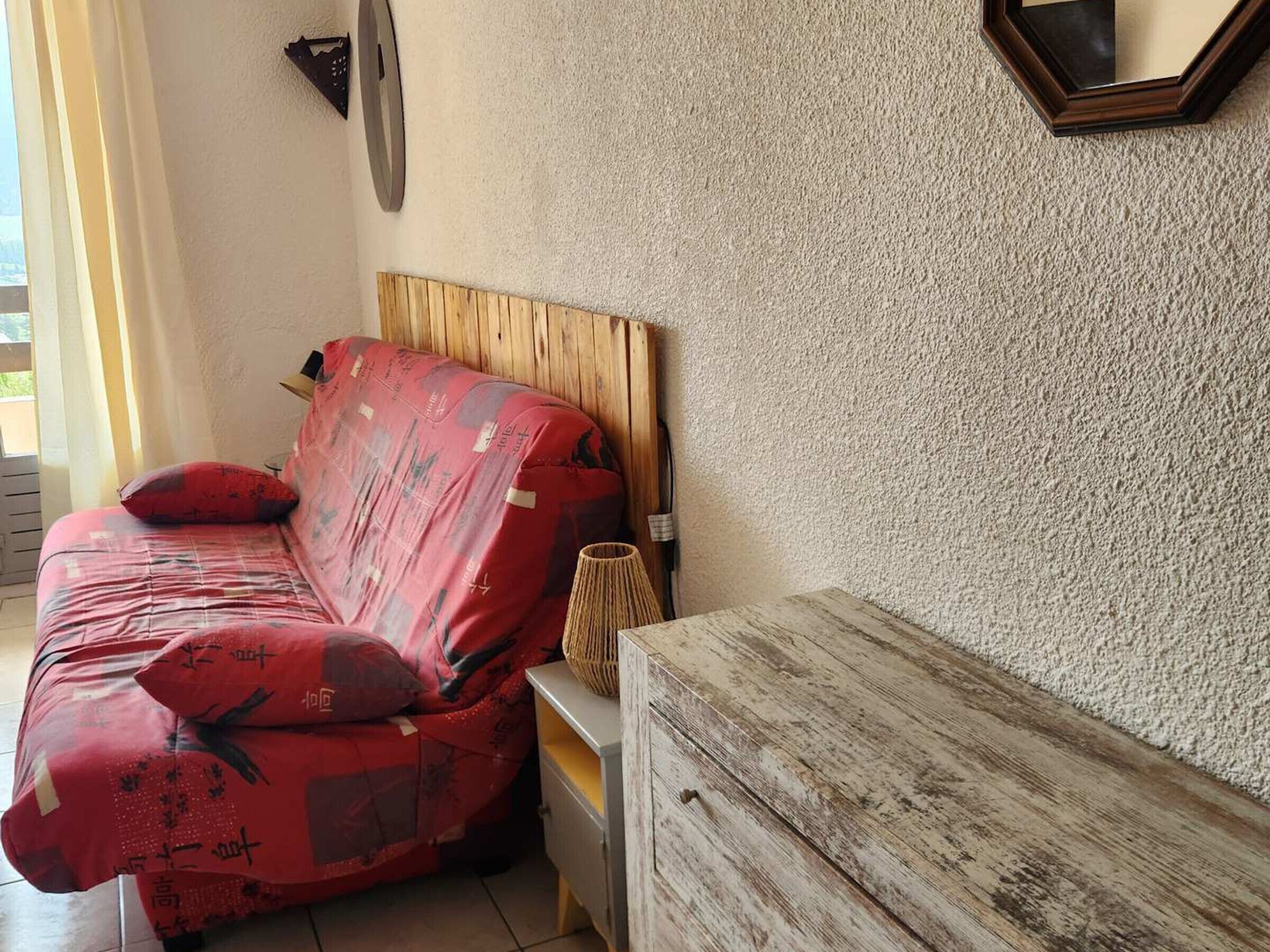 A room with a wooden headboard bed, a small bedside table with a lamp, and a plain wall.
