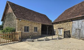 Ein Steinhaus mit Ziegeldach, ein von Zäunen umgebenes Gebiet mit Topfpflanzen und ein Holztor.