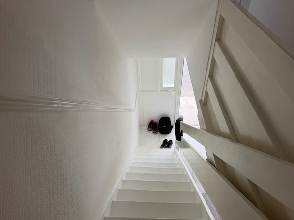 Een witte trap met een leuning, die uitkomt op een plateau waar schoenen en een rugzak staan.