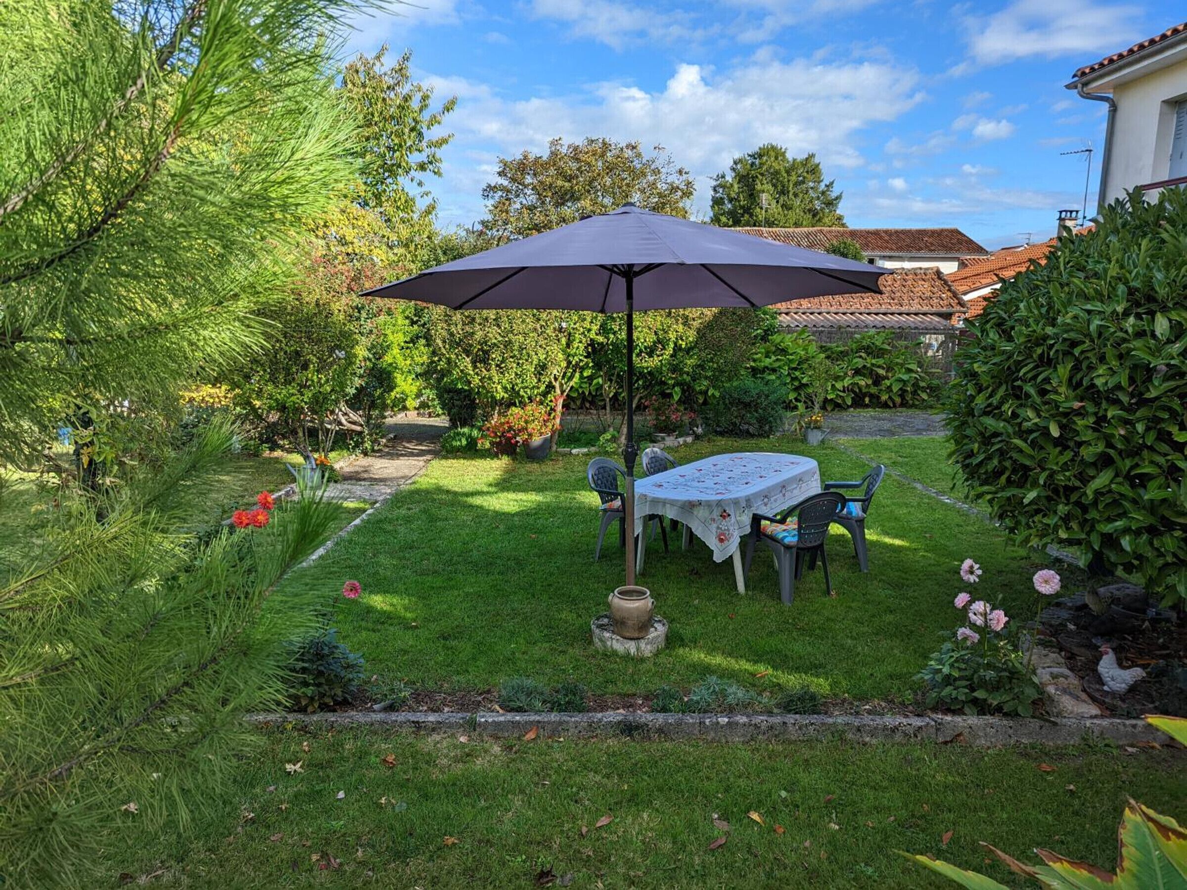 Un giardino con tavolo e sedie sotto un ombrellone grande, circondato da vegetazione e una casa sullo sfondo.