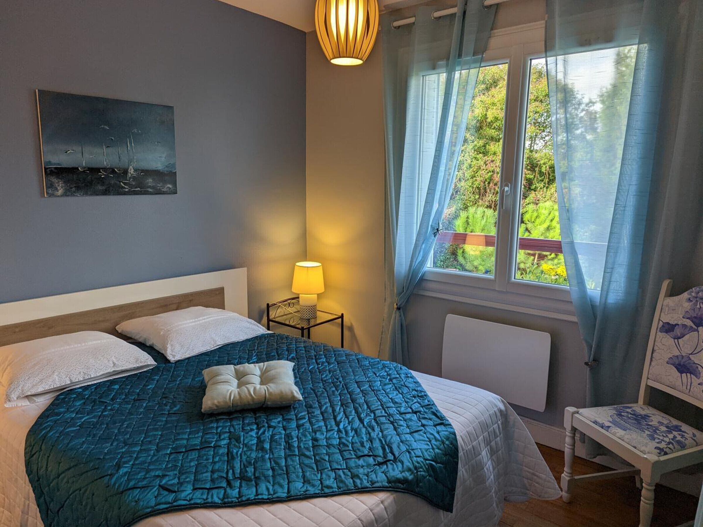 Un dormitorio con una cama, una lámpara de noche, una silla, un ventanal con cortinas y un cuadro en la pared.