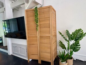 Ein hoher, hölzerner Raumteiler mit horizontalen Lamellen, eine kleine, grüne Pflanze darauf und eine große Monstera-Pflanze daneben.