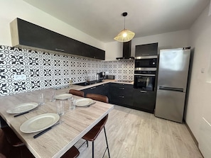 A modern kitchen with dark cabinets, a wooden dining table, and a built-in oven. - Bright apartment near the sea - 4 guests (La Barre-de-Monts)